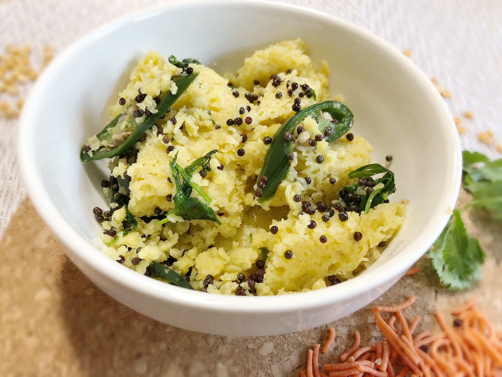 a bowl with khaman, mustard seeds, jalapenos, and cilantro as toppings. Surrounded by sev and extra garnished cilantro