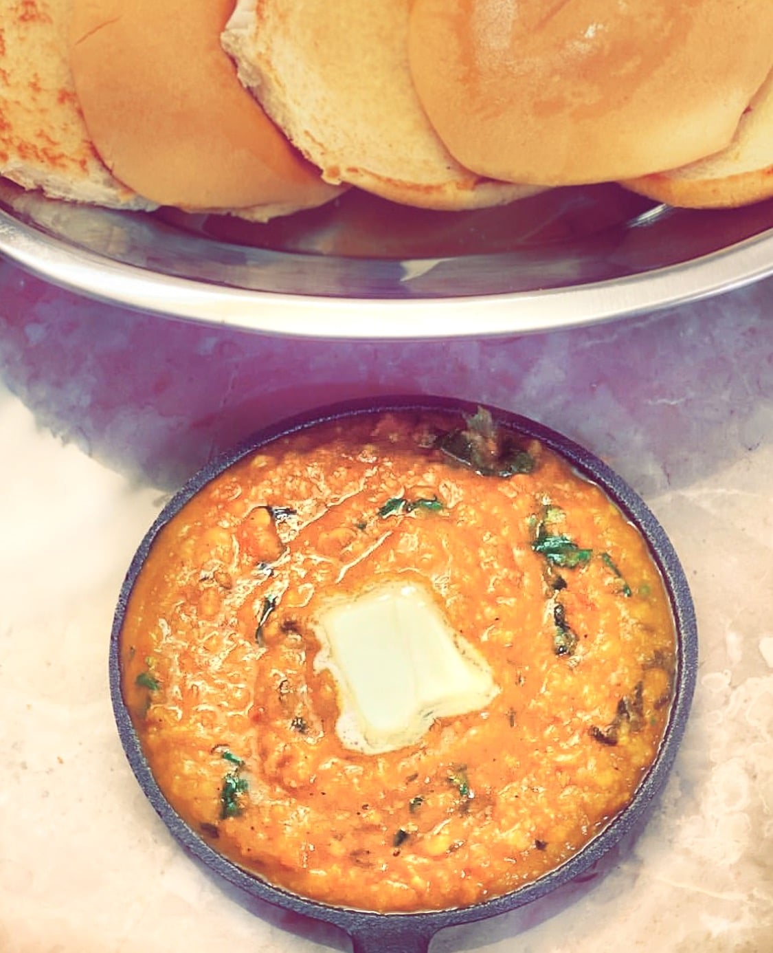 A cast iron pan with pav bhaji and butter on top with a layer of 5 grilled breads above it in a metal plate