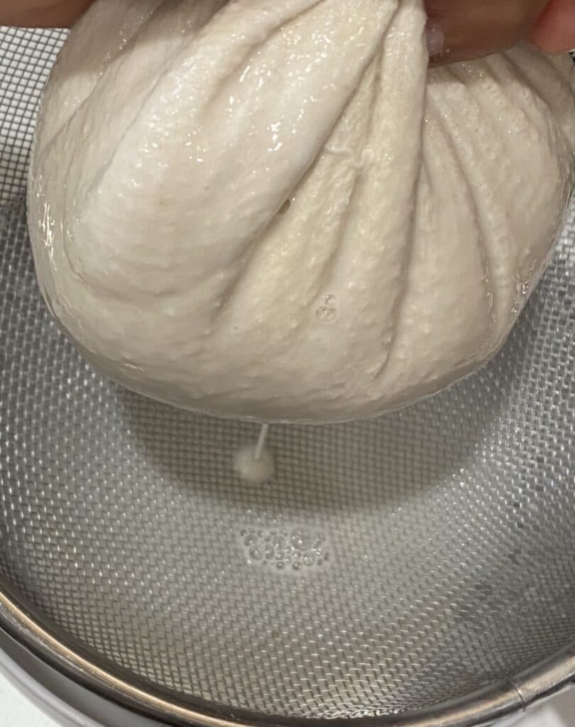 straining oatmilk out of a cheesecloth into a strainer
